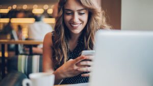 a woman seem to be laughing having mobile phone in her hand in front of laptop