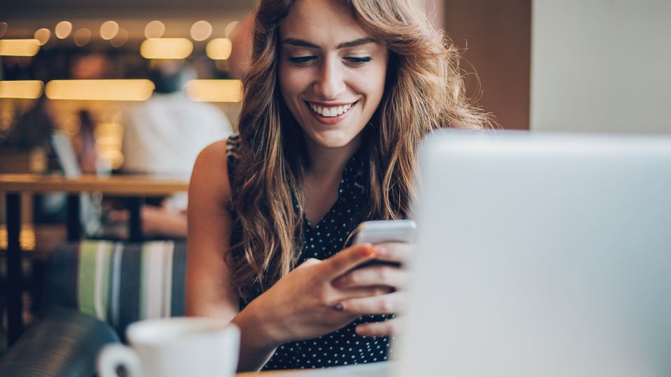 a woman seem to be laughing having mobile phone in her hand in front of laptop